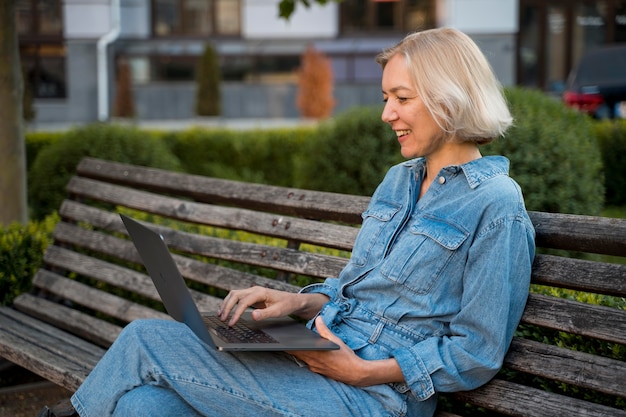 Foto vista lateral de uma mulher mais velha ao ar livre em um banco com um laptop