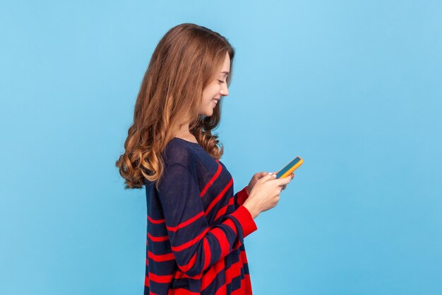 Vista lateral de uma mulher otimista sorridente em pé com telefone inteligente nas mãos, respondendo a mensagem romântica do namorado, vestindo suéter casual listrado, tiro de estúdio interior isolado em fundo azul