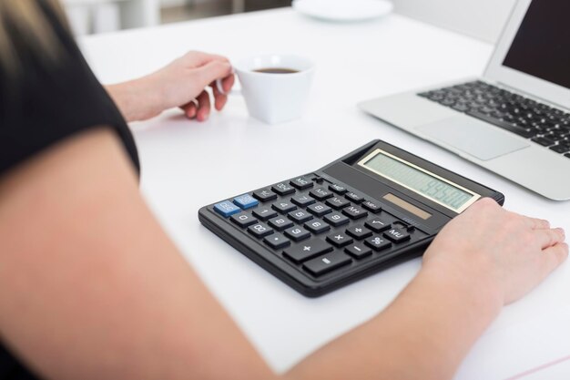 Vista lateral de uma mulher segurando uma xícara de café e usando sua calculadora. Há um laptop na mesa. Conceito de trabalho do contador