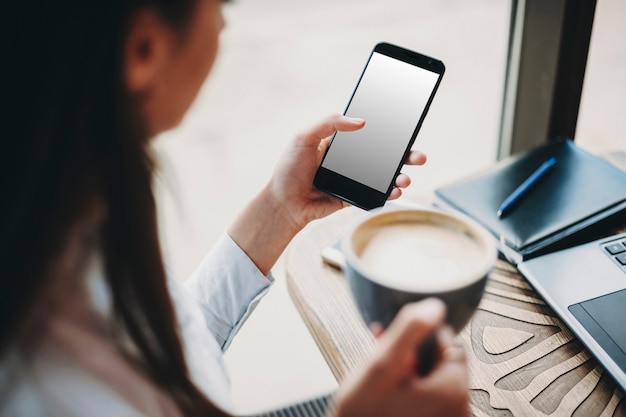 Vista lateral de uma mulher sentada em uma cafeteria com uma xícara de café na mão enquanto está em outra usando um smartphone. Conceito de banco online.