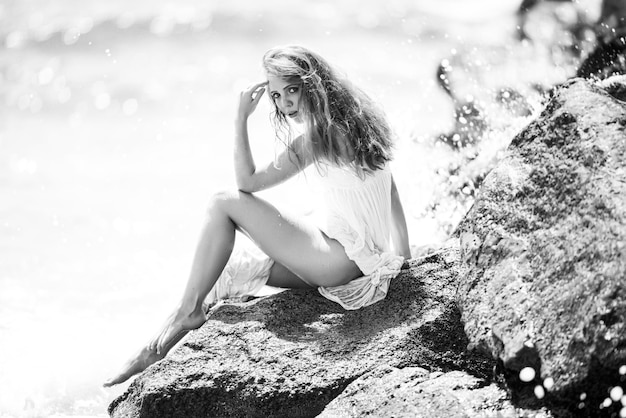 Vista lateral de uma mulher serena sentada na praia rochosa arenosa contra ondas azuis atraente jovem mulher wea