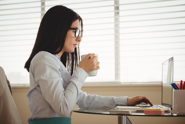 Vista lateral de uma mulher tomando café sentada ao lado do laptop