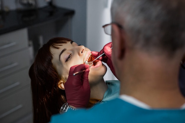 Vista lateral del dentista médico senior masculino en la cirugía del dentista con operación dental para paciente joven en clínica de estomatología moderna. Centrarse en la cara de mujer