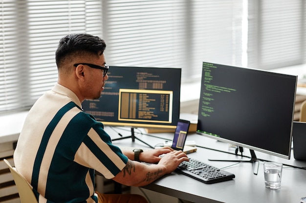 Vista lateral del desarrollador de ti asiático escribiendo en el teclado con código de programación en la pantalla de la computadora mientras wo