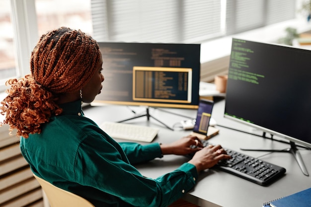 Vista lateral de la desarrolladora de TI femenina escribiendo en el teclado con código de programación en la pantalla de la computadora mientras w