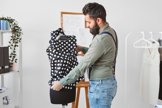 Vista lateral del diseñador de moda masculino barbudo que trabaja con la forma del vestido en el atelier