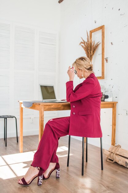 Vista lateral de una diseñadora segura de sí misma con cabello rubio mirando seriamente la pantalla de una laptop, trabajando en un nuevo proyecto, navegando por Internet. Retrato de cuerpo entero de una mujer de negocios con ropa elegante.