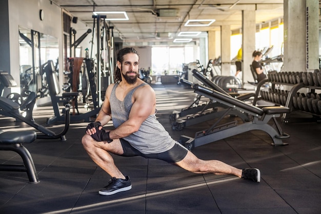 Vista lateral do atleta desportista adulto jovem bonito com cabelo longo encaracolado malhando na academia, agachando em um joelho, alongamento após o treino, fazendo exercícios para as pernas, de cócoras. interior, olhando para longe