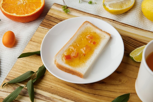 Vista lateral do café da manhã com fatia de pão manchada com geléia no prato e copo de suco de laranja fatia de limão com folhas na tábua e tripé de geléia e livro aberto sobre fundo de pano branco