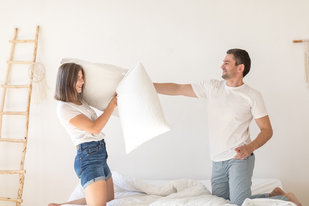 Vista lateral do casual casal brincalhão se divertindo e travesseiro lutando enquanto está sentado na cama na sala moderna, com paredes brancas