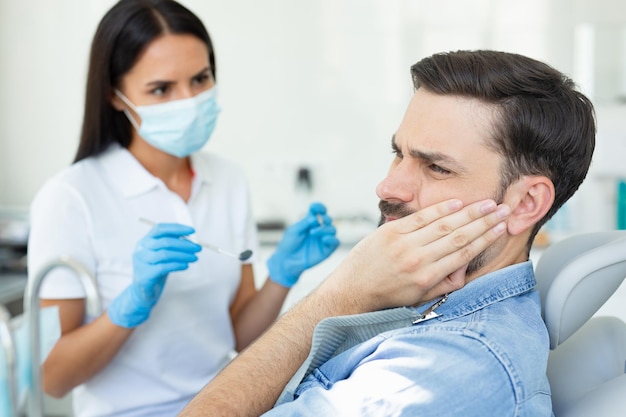 Vista lateral do cliente caucasiano tocando o rosto enquanto tem dor de dente perto do dentista no hospital