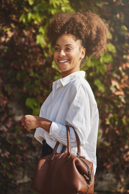 Vista lateral do conteúdo empresário feminino negro na camisa branca com bolsa sorrindo e olhando para longe em pé no parque da cidade num dia de verão