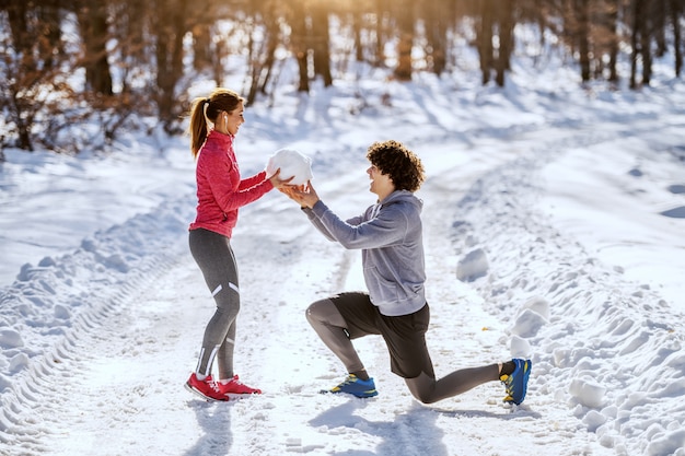 Vista lateral do engraçado homem caucasiano ajoelhado na frente de sua namorada e dando-lhe grande bola de neve. Ambos estão vestidos com roupas esportivas. Inverno.
