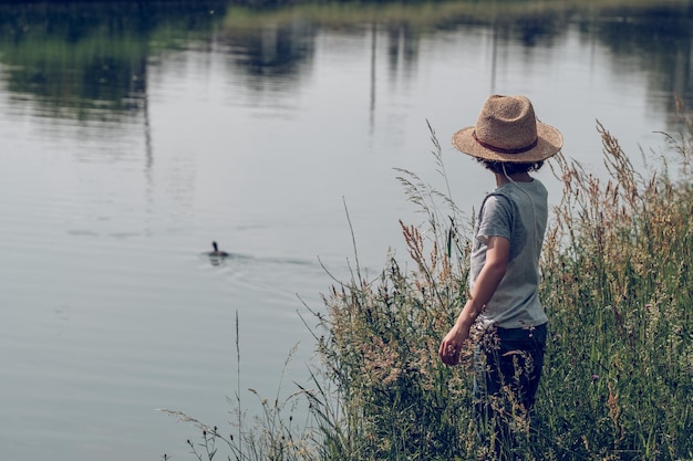 Vista lateral do garoto irreconhecível com chapéu de palha em pé entre a grama verde na margem do canal e olhando para o pato na água em dia de verão