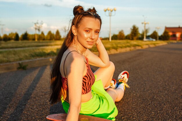 Vista lateral do hipster feminino sentado no skate na estrada no verão ao pôr do sol e olhando para a câmera