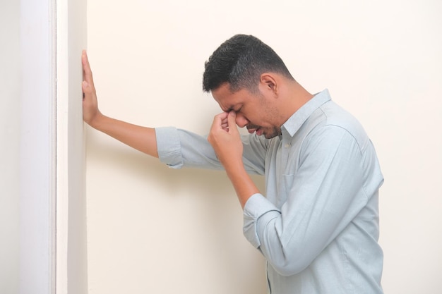 Vista lateral do homem asiático adulto mostrando expressão triste com uma mão segurando na parede