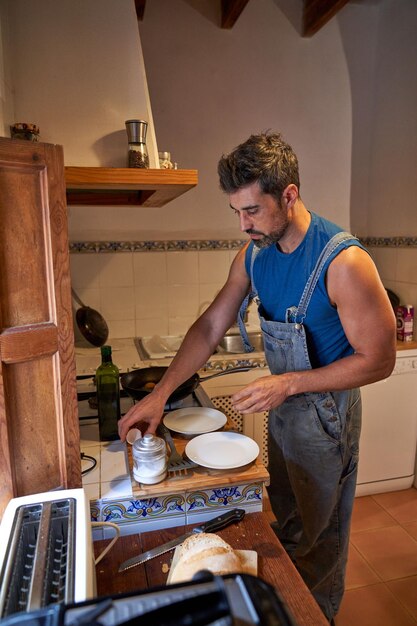 Vista lateral do homem barbudo adulto concentrado no avental em pé perto do balcão de madeira enquanto cozinhava refeição para dois no fogão a gás na cozinha pela manhã