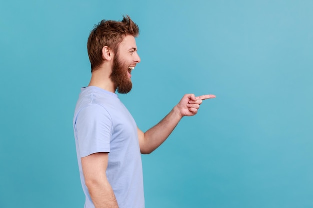 Vista lateral do homem barbudo satisfeito positivo apontando de lado mostrando espaço de cópia em branco para texto comercial de apresentação de ideia sorrindo alegremente tiro de estúdio interno isolado em fundo azul