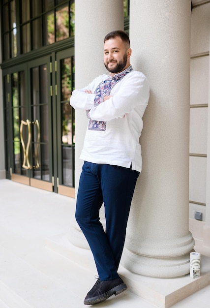 Vista lateral do homem bonito com barba e bigode vestido com camisa étnica cruzando as mãos e olhando para a câmera enquanto se apoia na coluna de arquitetura do edifício moderno ao ar livre