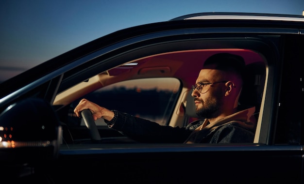 Vista lateral do homem bonito sem barba em roupas da moda que andam de automóvel