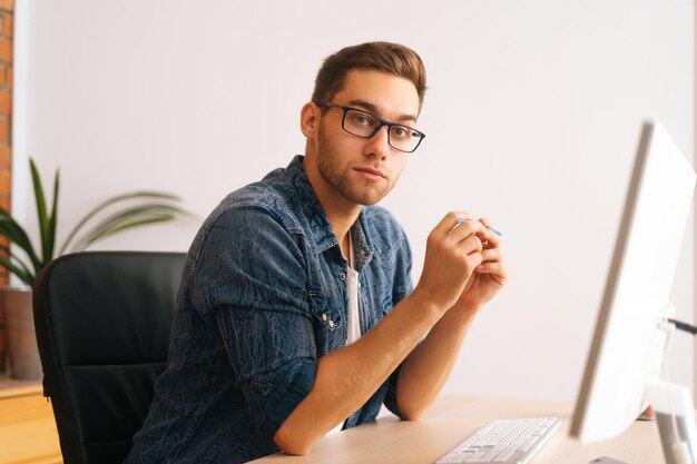 Vista lateral do homem de designer freelance jovem bonito sério em óculos elegantes, trabalhando no computador desktop, segurando a caneta nas mãos, sentado na mesa no escritório em casa, olhando para a câmera.