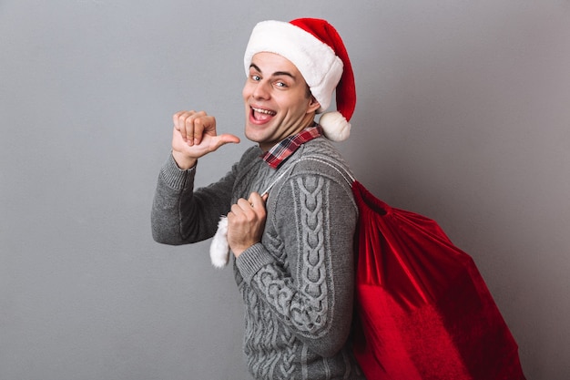 Vista lateral do homem feliz com suéter e chapéu de Natal segurando uma sacola com presentes, apontando para trás e olhando