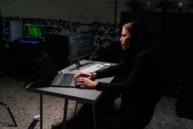 Vista lateral do homem hacker perigoso em moletom hackeando site online envolvendo hacking em sistemas de segurança usando laptop de digitação