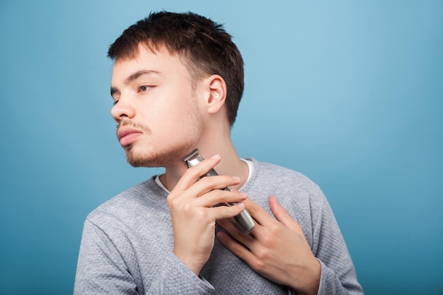 Vista lateral do homem moreno concentrado com cerdas no suéter casual raspando o pescoço atento e cuidadoso usando o aparador para moldar o estúdio de beleza e higiene da barba isolado no fundo azul