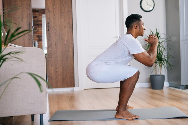 Vista lateral do homem musculoso muscular afro-americano exercitando e fazendo agachamentos durante o exercício no quarto doméstico brilhante. Conceito de treinamento esportivo no ginásio em casa.