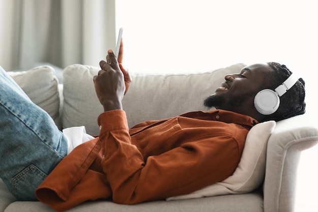 Vista lateral do homem negro ouvindo música no smartphone online descansando no sofá em casa espaço livre