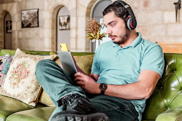 Vista lateral do homem sério com fones de ouvido usando um tablet e celular sentado em um sofá em casa