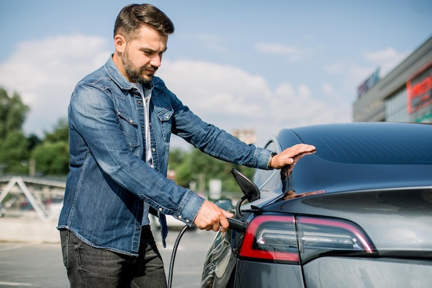 Vista lateral do jovem barbudo bonito em camisa jeans conectando o fio na tomada do carro para carregar seu novo carro elétrico moderno de luxo na estação de carregamento ao ar livre na cidade