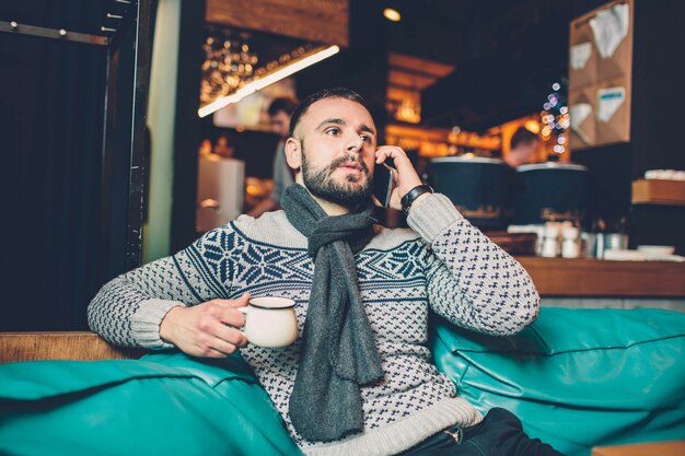 Vista lateral do jovem barbudo, vestido incasual desgaste, sentado na mesa de madeira redonda no café com interior moderno e está segurando o smartphone.