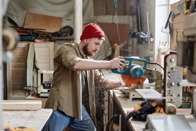 Vista lateral do jovem carpinteiro caucasiano moderno cortando prancha de madeira com serra de mitra no trabalho de marcenaria
