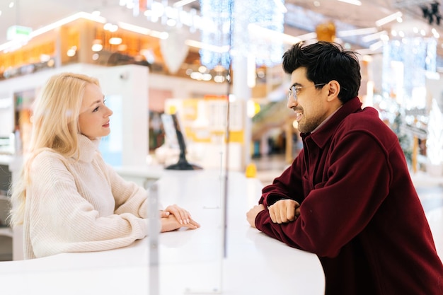 Vista lateral do jovem casal feliz de homem barbudo elegante em óculos e uma linda mulher loira conversando através da divisória de vidro em pé no corredor do shopping