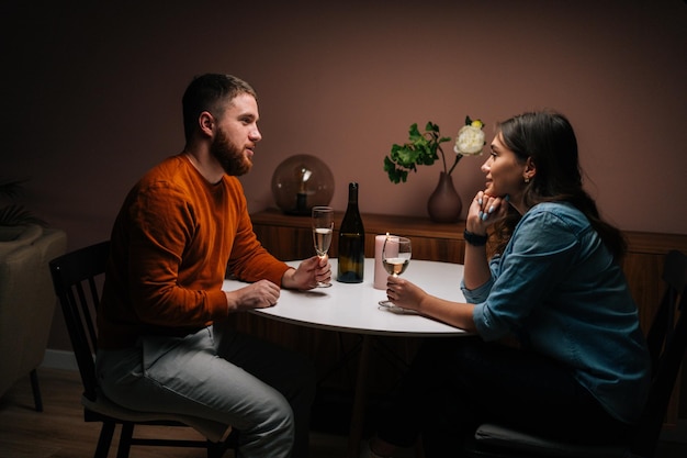 Vista lateral do jovem casal feliz gostando de conversar e sentar juntos à mesa de jantar com velas no quarto escuro aconchegante