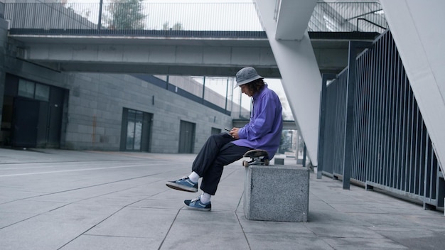 Vista lateral do jovem desportivo sentado no skate perto do estádio da cidade com telefone celular Hipster elegante digitando na tela do celular ao ar livre Cara de patinador focado usando smartphone do lado de fora