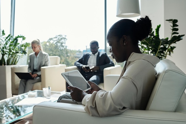 Vista lateral do jovem trabalhador de escritório feminino sério com tablet sentado no sofá