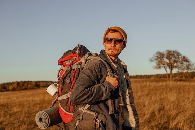 Foto vista lateral do jovem viajante masculino com mochila de caminhada em roupa hipster em pé no campo ao pôr do sol