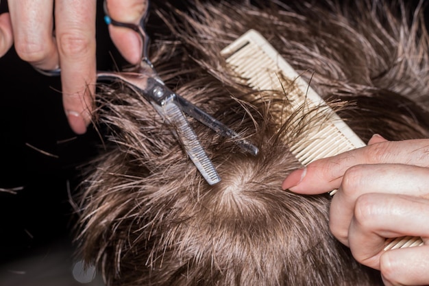 Vista lateral do menino bonitinho cortando o cabelo pelo cabeleireiro na barbearia usando pente e tesoura de aliciamento. Closeup, o conceito de moda e beleza.