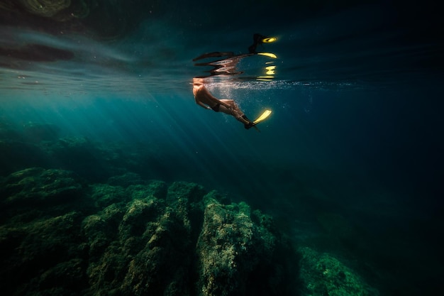Vista lateral do mergulhador anônimo em flipper nadando sob a água azul do mar profundo perto de recifes de corais ásperos