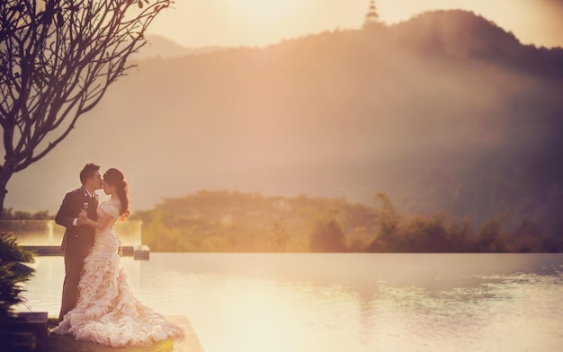 Foto vista lateral do noivo beijando a noiva enquanto está ao lado do lago durante o pôr do sol