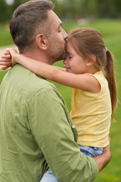 Foto vista lateral do pai agradecido segurando e beijando sua filhinha enquanto passam um tempo juntos ao ar livre