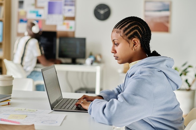 Vista lateral do programador com penteado elegante trabalhando no laptop sentado em seu local de trabalho no escritório