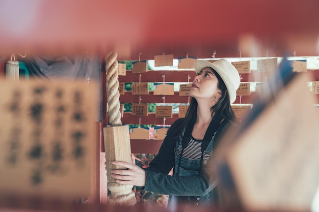 vista lateral do santuário de madeira da mulher japonesa puxando a mão tocando a corda shimenawa fazer sino suzu no histórico templo de madeira. menina com chapéu carregando câmera rezando em shitennoji xintoísmo osaka japão