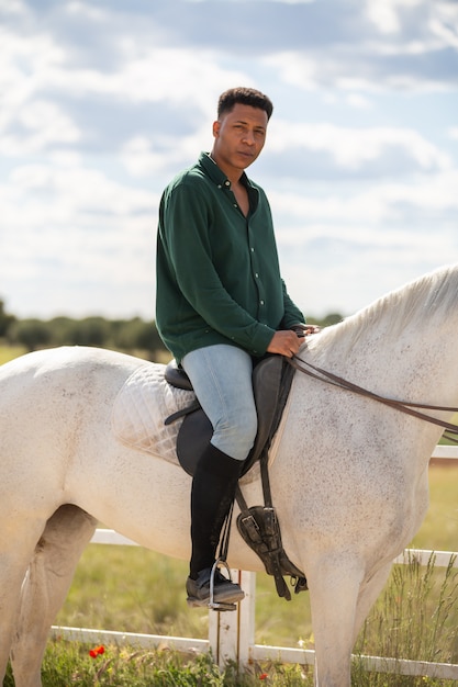 Vista lateral do sorridente jovem afro-americano masculino olhando para a câmera enquanto está sentado na traseira do cavalo branco