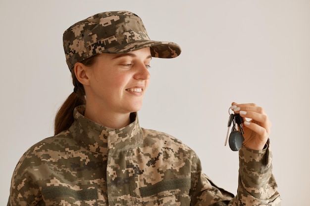 Vista lateral do sorridente soldado feminino vestindo traje militar, posando no interior da sala de luz, em pé com a chave nas mãos, comprando uma nova compra, tendo o orgulho de comprar a casa.