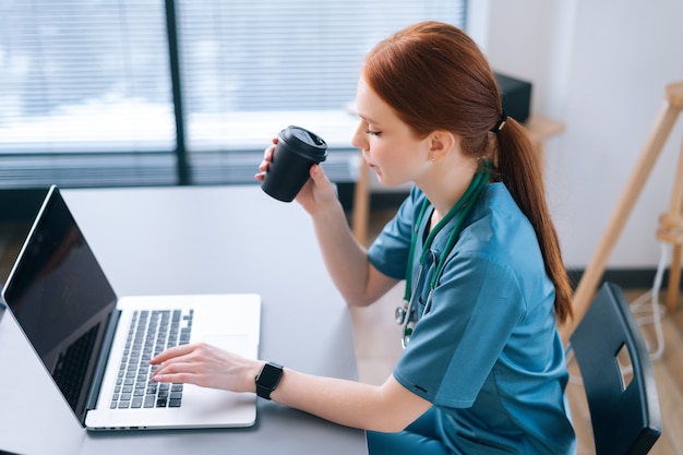 Vista lateral de una doctora seria que brinda una consulta en línea a distancia al paciente a través de una computadora portátil y tomando café de la taza