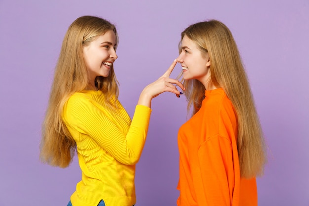 Vista lateral de dos hermanas gemelas bastante rubias niñas en ropa colorida poniendo el dedo en la nariz divirtiéndose aislado en la pared azul violeta pastel. Concepto de estilo de vida familiar de personas.