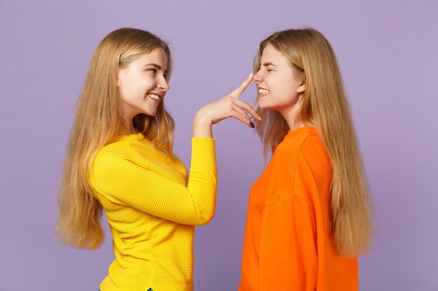 Vista lateral de dos jóvenes hermanas gemelas rubias niñas en ropa colorida poniendo el dedo en la nariz, divirtiéndose aislado en la pared azul violeta pastel. Concepto de estilo de vida familiar de personas.